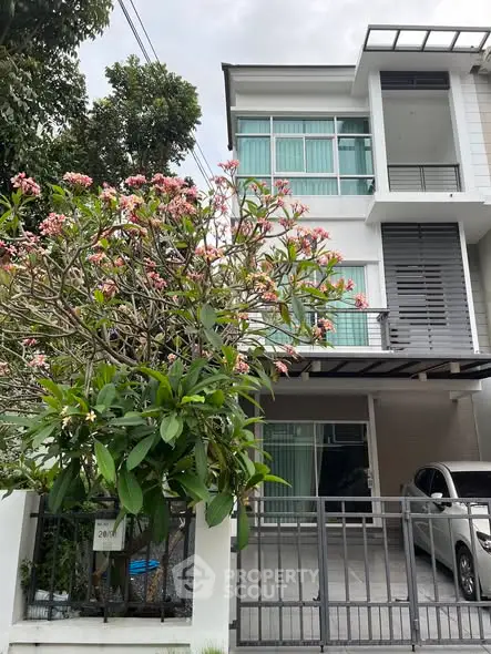 Modern three-story house with a carport and lush flowering tree in front, showcasing contemporary architecture.