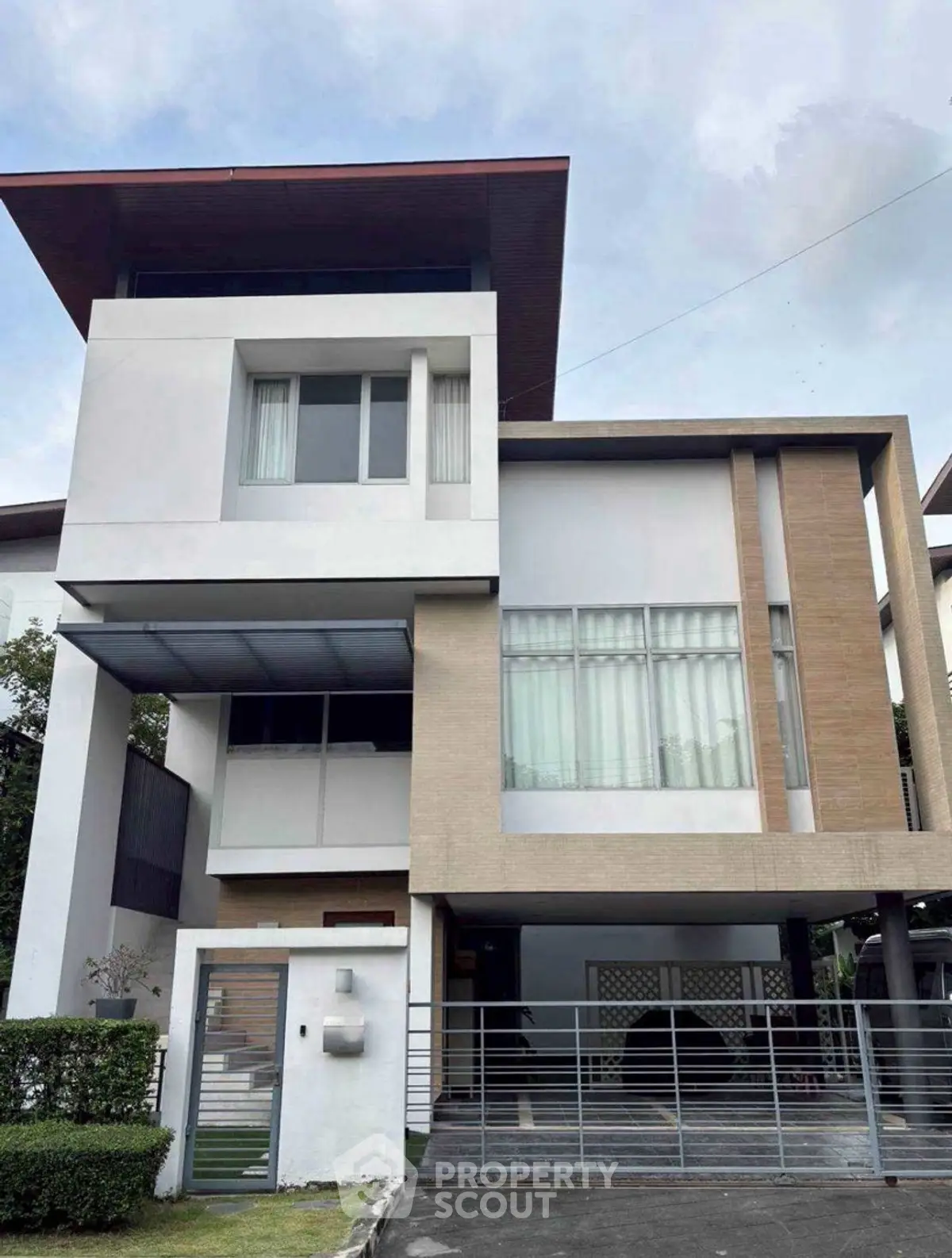 Modern three-story house with sleek design and large windows, featuring a gated driveway and lush greenery.