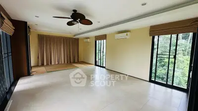 Spacious living room with large windows and ceiling fan, offering serene views of lush greenery.