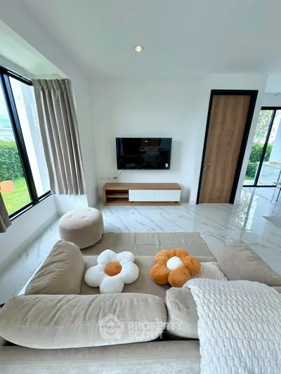 Modern living room with stylish beige sofa and wall-mounted TV, featuring large windows for natural light.
