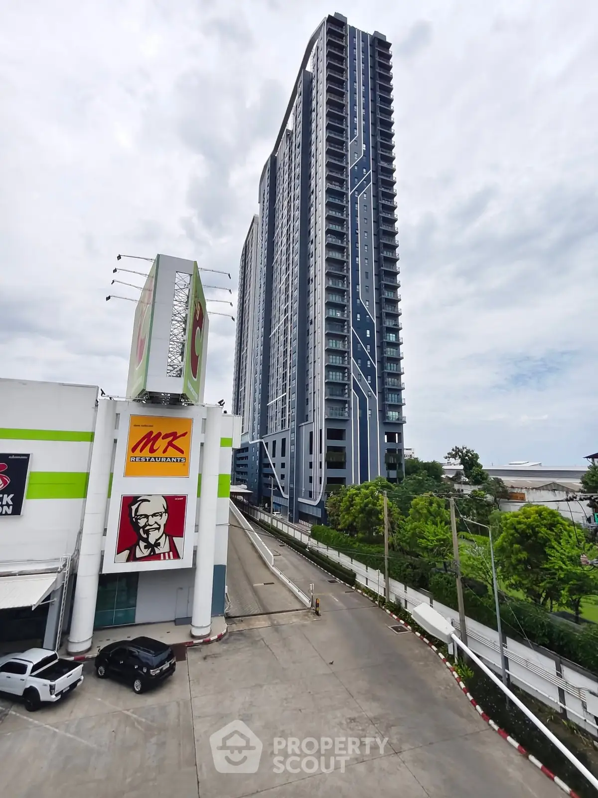 Modern high-rise building next to commercial area with ample parking space and lush greenery.