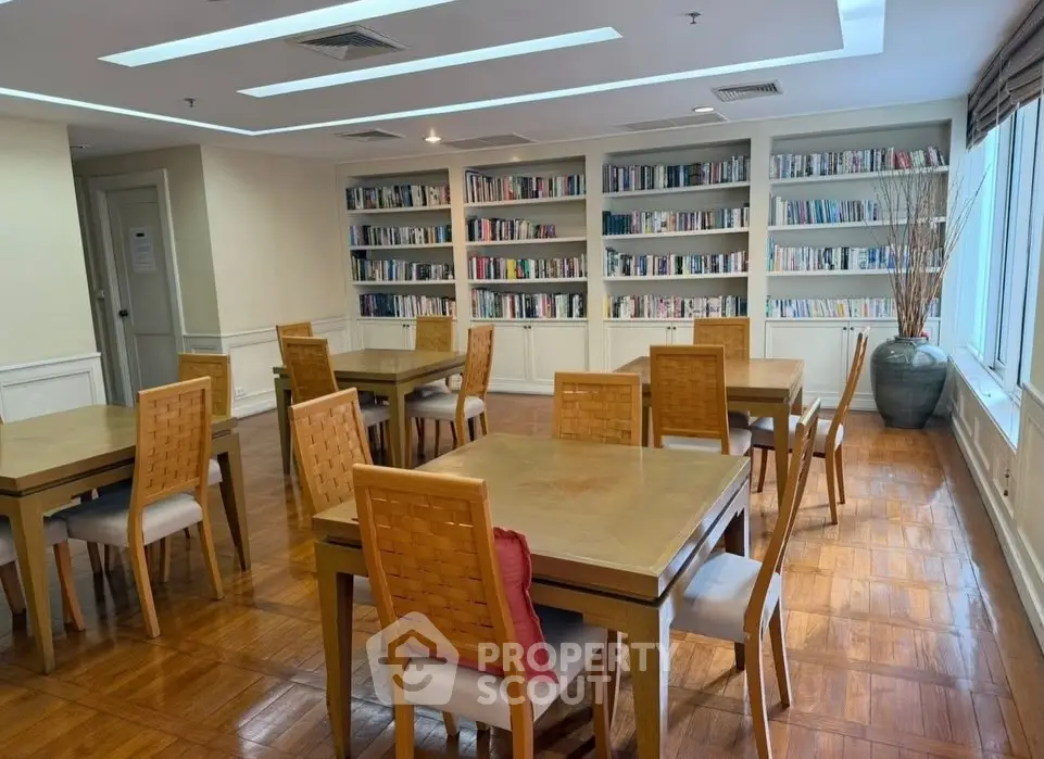 Spacious library with wooden tables and chairs, surrounded by bookshelves and natural light.