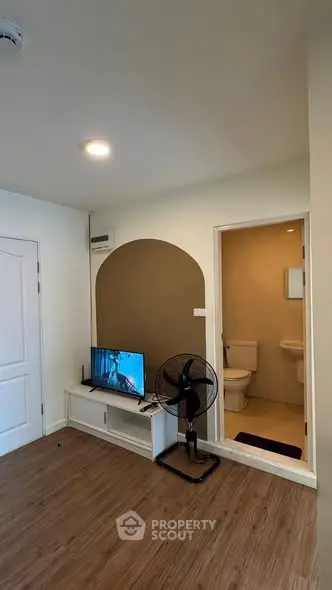 Cozy living room with TV and fan, adjacent to a compact bathroom, featuring wooden flooring and neutral tones.