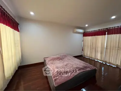 Spacious bedroom with elegant wooden flooring and cozy bed, featuring large windows with red curtains.