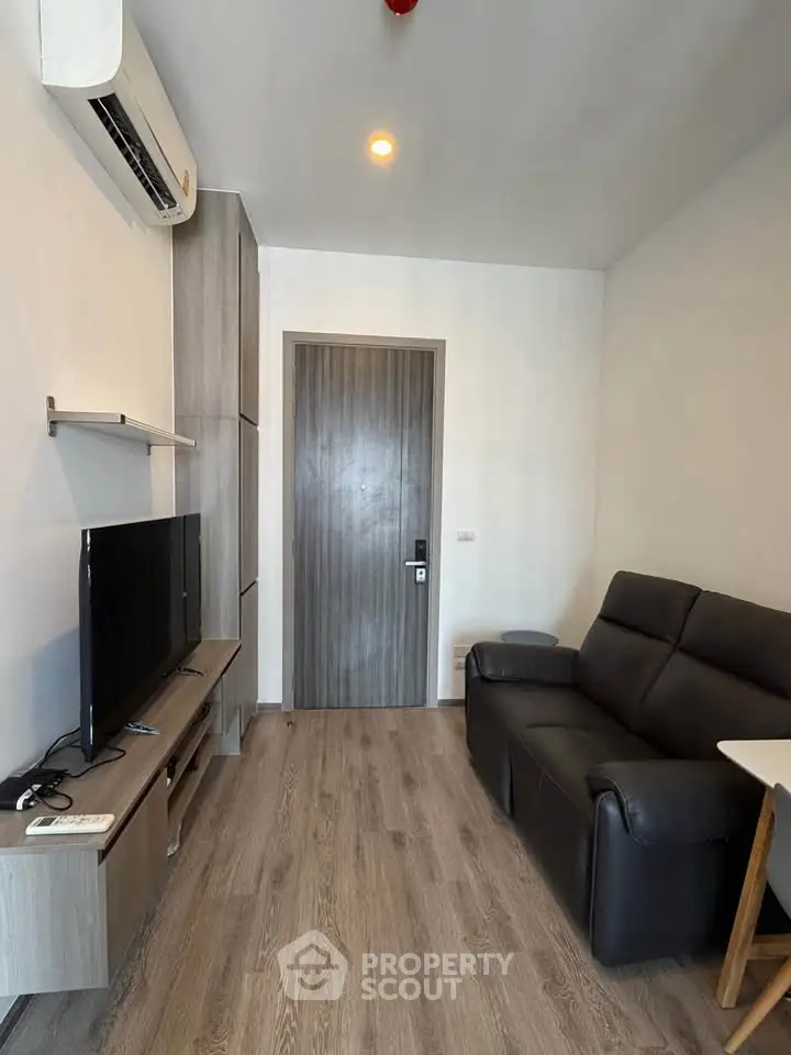 Modern living room with sleek furniture and wooden flooring, featuring a flat-screen TV and air conditioning.