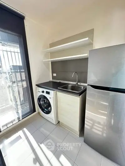 Modern compact kitchen with washing machine and fridge, natural light from large window.