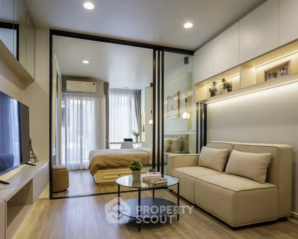 Modern living room with glass partition leading to cozy bedroom, featuring sleek furniture and ambient lighting.