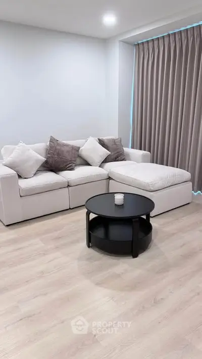 Modern living room with stylish white sectional sofa and round black coffee table on light wood flooring.