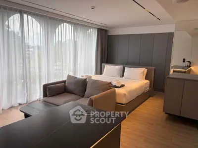 Modern studio apartment with cozy bed, stylish sofa, and large windows offering natural light.
