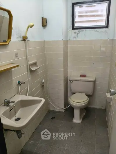 Compact bathroom with beige tiles, featuring a sink, toilet, and showerhead under a small window.