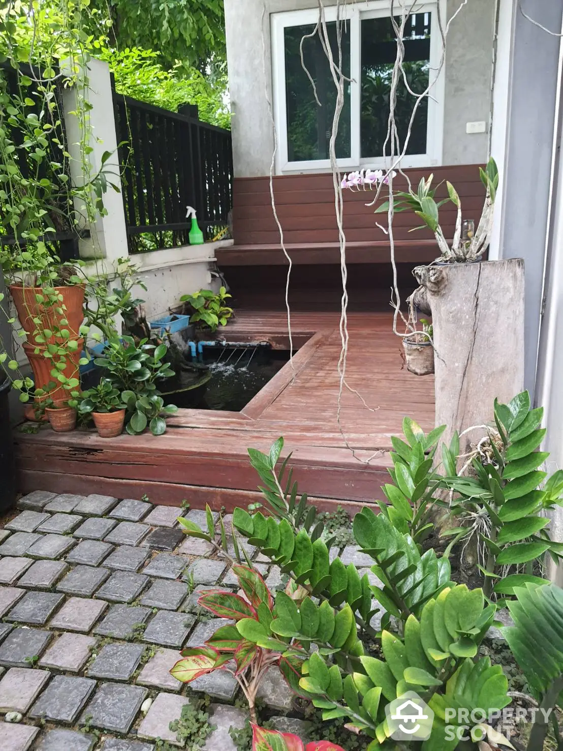 Charming home entrance with a wooden deck and lush greenery, featuring a cozy porch surrounded by potted plants and a serene pond, creating a welcoming atmosphere.