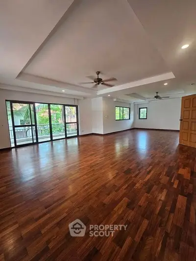 Spacious living room with wooden flooring and ceiling fans, featuring large windows for natural light.