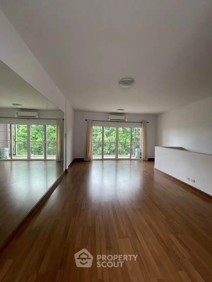 Spacious living room with large windows and wooden flooring, offering a serene view of lush greenery.