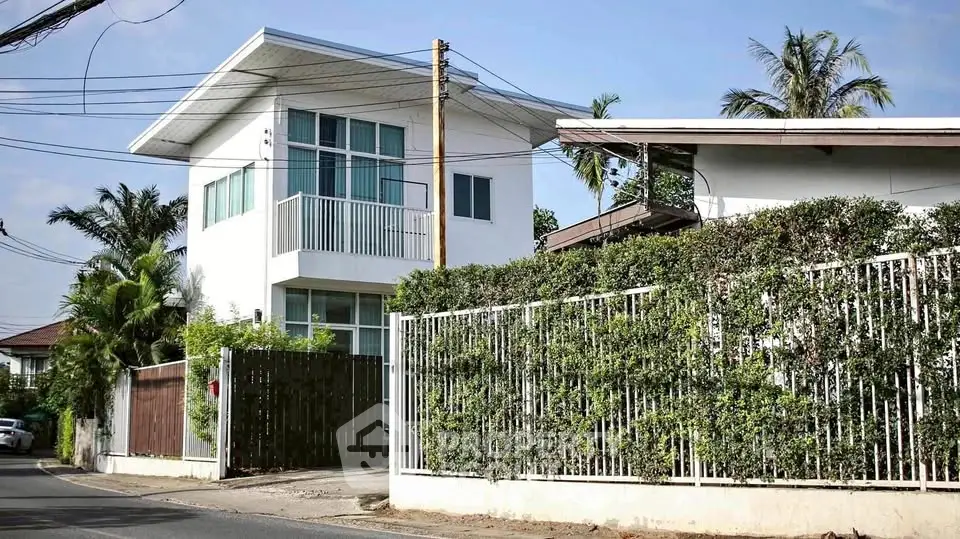 Modern two-story house with lush greenery and white fence in suburban setting.