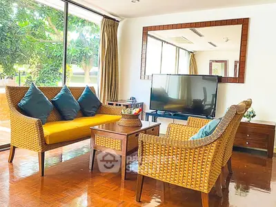 Charming living room with wicker furniture and large windows offering lush garden views.