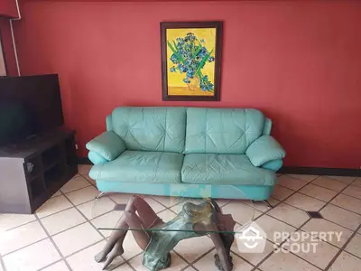 Vibrant living room with a striking red wall, teal sofa, and unique glass coffee table, complemented by a colorful artwork and tiled flooring.