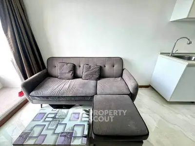 Modern living room with sleek gray sofa and glass coffee table, adjacent to compact kitchen area.