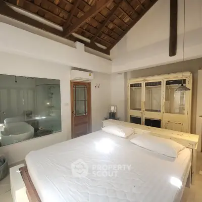 Charming bedroom with rustic wooden ceiling and large window overlooking a modern bathroom with a bathtub.