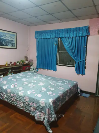 Cozy bedroom with floral bedding and blue curtains, featuring a wooden floor and decorative wall art.