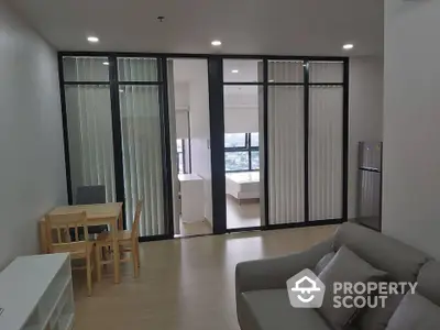 Modern open-plan living room with dining area and view into bedroom, featuring sleek furniture and large windows.