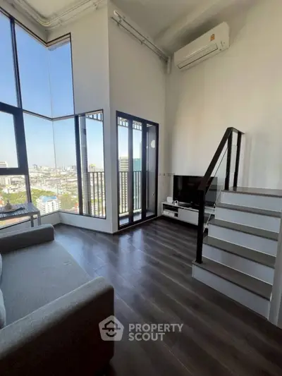Modern loft apartment with high ceilings, large windows, and city view, featuring sleek staircase and cozy living area.