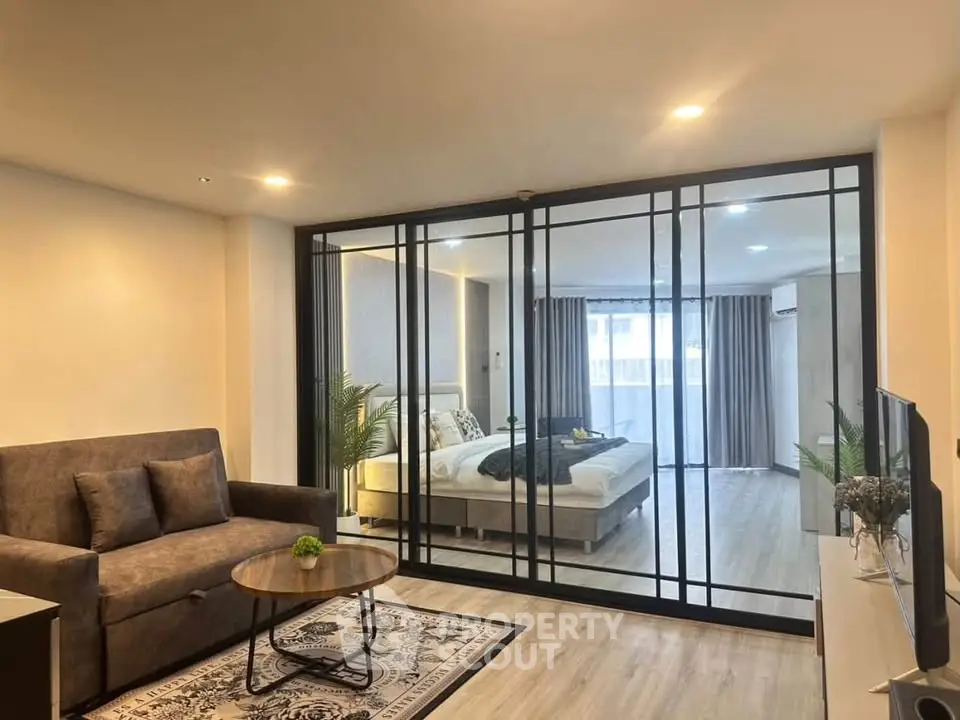 Modern living room with glass partition leading to a stylish bedroom, featuring cozy furniture and elegant decor.