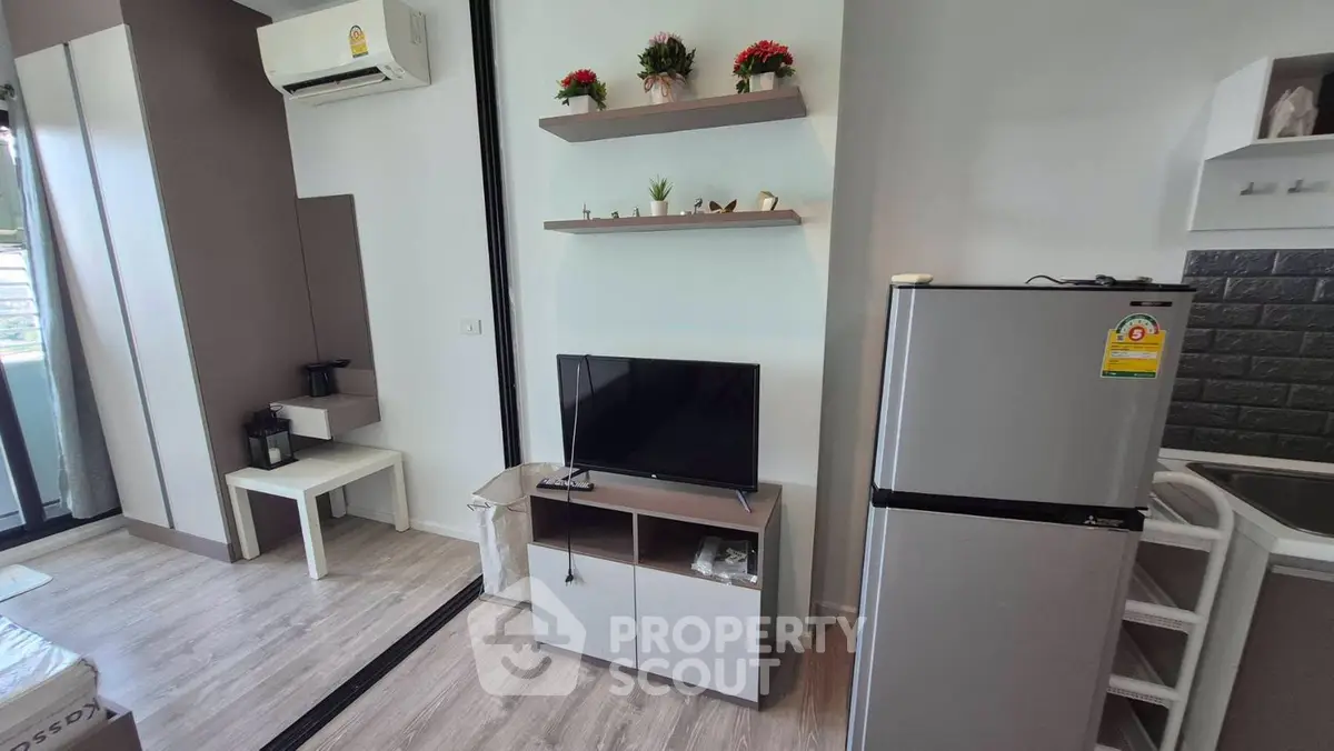 Modern living room with sleek decor and compact kitchen area, featuring a flat-screen TV and stainless steel fridge.