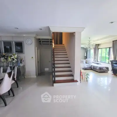 Spacious living room with elegant staircase and dining area, featuring large windows and modern decor.