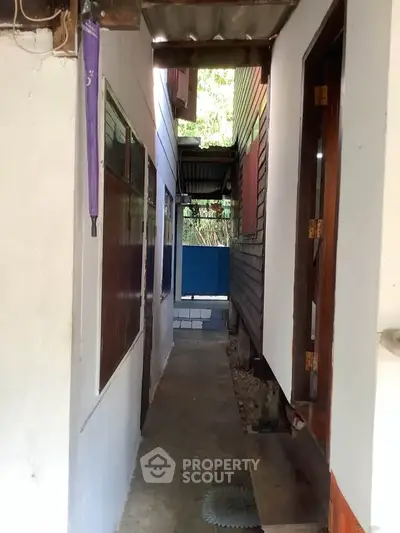 Narrow outdoor corridor between two buildings with wooden and white walls, leading to a small open area.