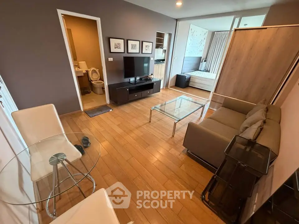 Modern living room with wooden flooring, glass table, and cozy seating area leading to a bedroom.