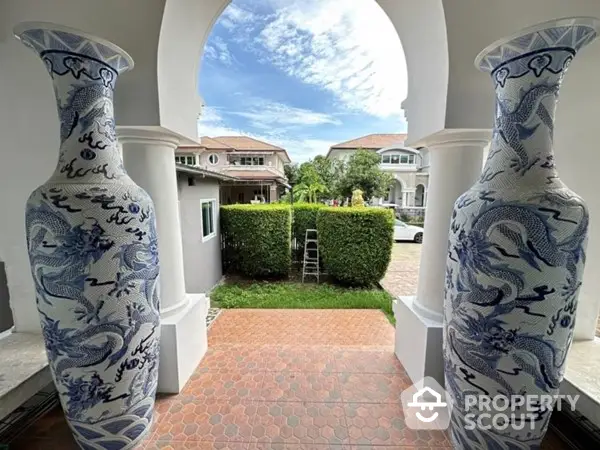 Elegant estate entrance framed by two large traditional blue and white porcelain vases, showcasing a luxurious approach to a manicured garden and grand villas.