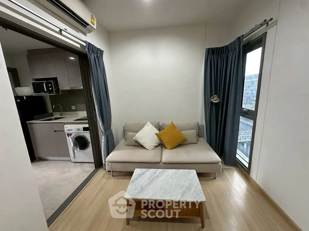Modern living room with open kitchen and washing machine, featuring cozy sofa and large windows.