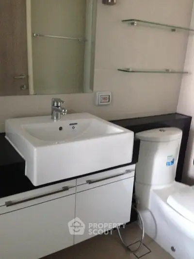 Modern bathroom with sleek sink and toilet, featuring glass shelves and contemporary design.