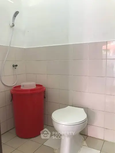 Compact bathroom with white toilet and red bucket, featuring tiled walls and a shower head.