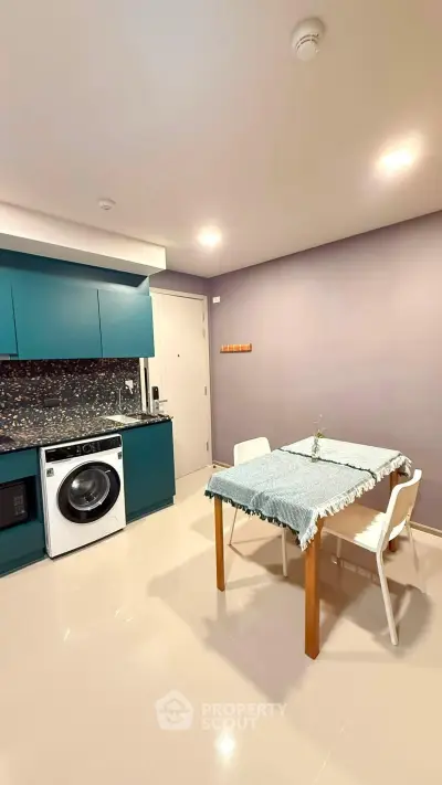 Modern kitchen with teal cabinets, washing machine, and dining area in a stylish apartment.