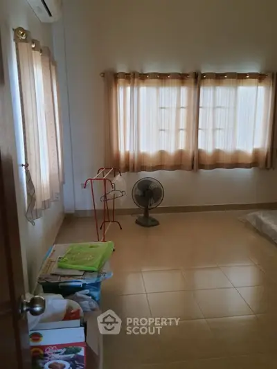 Spacious and well-lit living room with large windows draped in golden curtains, tiled flooring, and a vintage standing fan, awaiting personal touches.