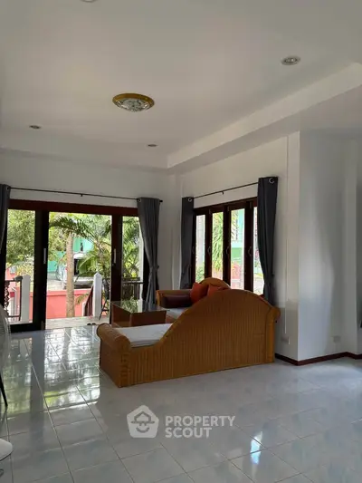 Spacious living room with large windows and garden view, featuring elegant wicker furniture and bright natural light.