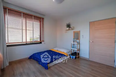 Cozy bedroom with wooden flooring and large window with blinds, featuring a minimalist design and natural light.