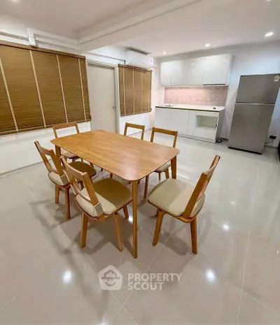 Spacious dining area with modern wooden table and chairs, adjacent to sleek kitchen with fridge.