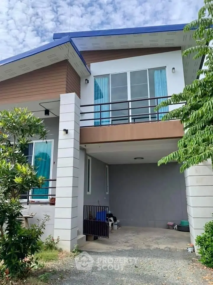 Modern two-story house with balcony and carport, surrounded by lush greenery.
