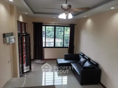 Spacious living room with modern black leather sofa and ceiling fan, featuring large window with garden view.