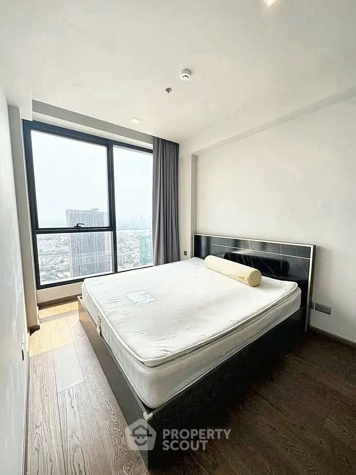 Modern bedroom with large window offering city views, featuring a sleek bed and wooden flooring.