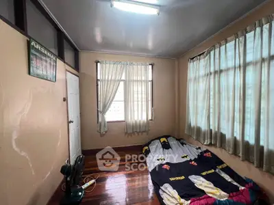 Cozy bedroom with wooden flooring and natural light, featuring a simple bed setup and fan.