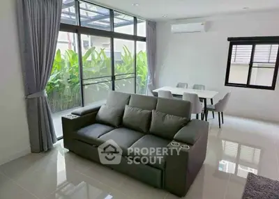 Modern living room with sleek sofa and dining area, featuring large windows and lush garden view.