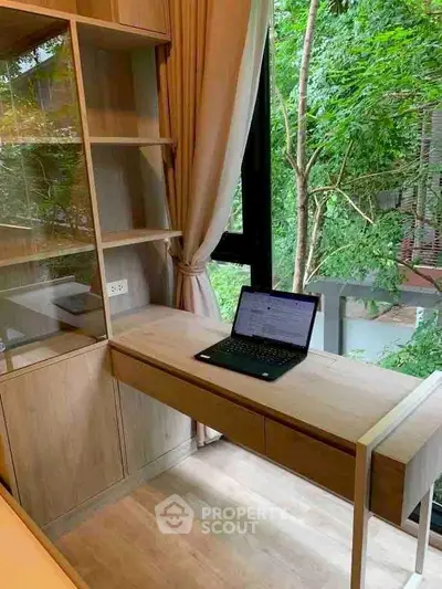 Modern study area with desk and laptop, overlooking lush greenery through large window.
