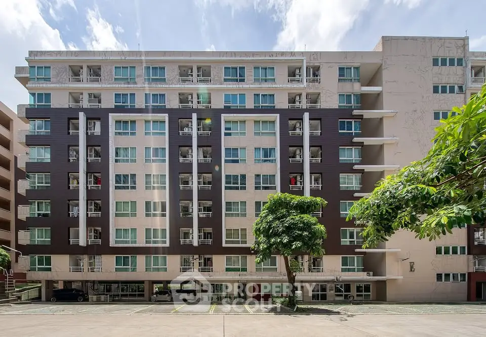 Modern mid-rise apartment building with balconies and parking area, set against a clear blue sky.
