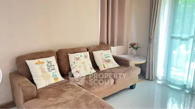 Cozy living room with brown sofa and decorative pillows, large window with curtains, and a side table with flowers.