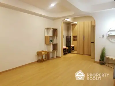 Spacious and well-lit living room with elegant curved ceiling design, wooden floors, and modern shelving units, leading to a cozy bedroom.