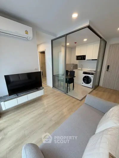 Modern living room with open kitchen and glass partition, featuring sleek furniture and appliances.