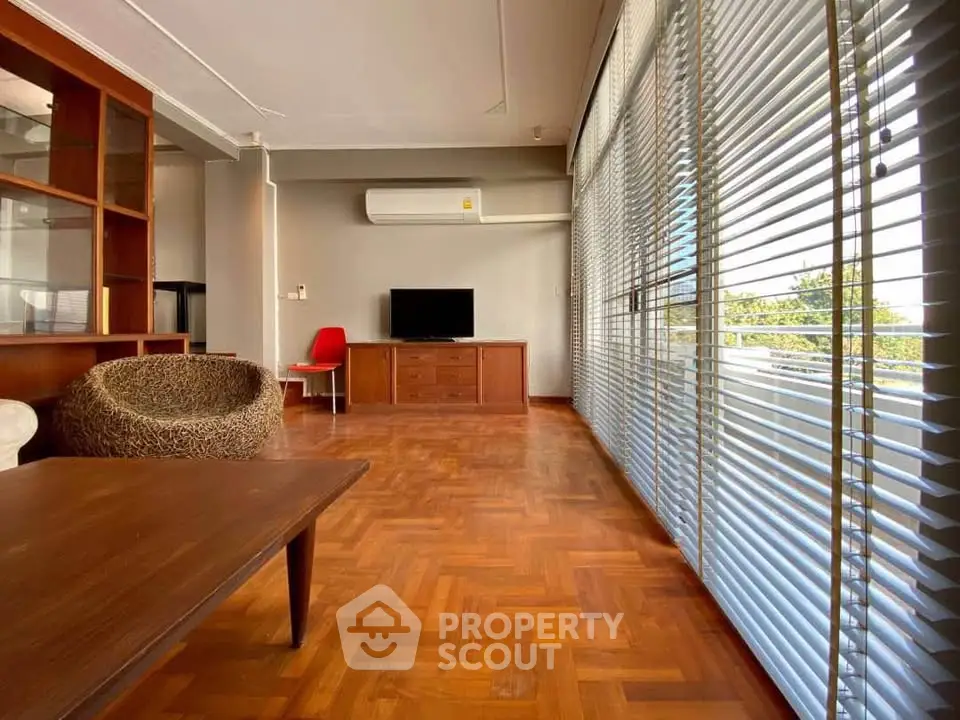Spacious living room with wooden flooring and large windows with blinds, featuring modern furniture.